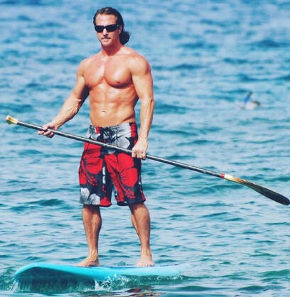 Scott Aloha stand-up paddleboarding on clear blue ocean water, wearing sunglasses and red board shorts